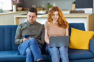 Handsome guy and his girlfriend using modern gadgets while sitting on couch at home.