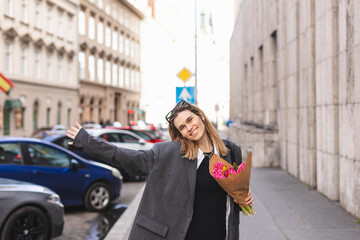 Fototapeta premium Positive girl hold pink flowers in the city center. Shot of amazing blonde haired lady with natural make up look happy, walking outdoors in grey jacket and hold pink bouquet in craft paper, open hands