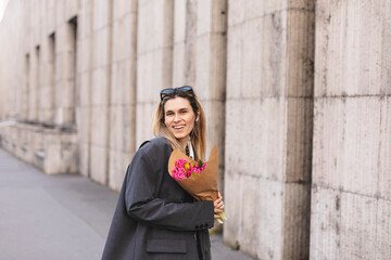 Positive girl hold pink flowers in the city center. Amazing blonde haired lady with natural make up look happy, walking outdoors in grey jacket and hold pink bouquet in craft paper, girl look happy.