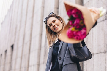 Positive girl hold pink flowers in camera. Close up shot of amazing blonde haired lady look happy, walking outdoors in grey jacket, sunglasses and hold pink bouquet in craft paper. Carefree, freedom.