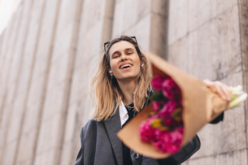 Positive girl hold pink flowers in camera. Close up shot of amazing blonde haired lady look happy, walking outdoors in grey jacket, sunglasses and hold pink bouquet in craft paper. Carefree, freedom.