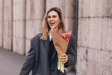 Positive girl hold pink flowers in the city center. Amazing blonde haired lady with natural make up look happy, walking outdoors in grey jacket and hold pink bouquet in craft paper, girl look happy.