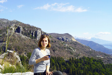 Naklejka premium Portrait of a female tourist on vacation - a smiling young woman sits with a hiking cup in her hand against the backdrop of a beautiful mountain landscape.