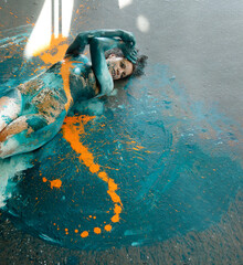expressive sexy naked woman elegant on the floor in turquoise blue and orange color abstractly painted bodypainting girl on the splashed ground, surrounded by sun rays, abstract body art
