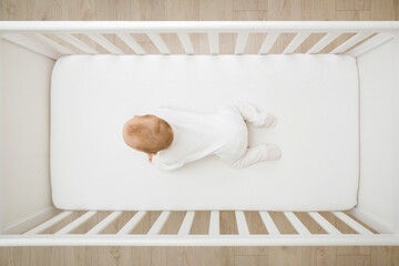 Fototapeta na wymiar Baby in white bodysuit try crawling on knee and arms on mattress in wooden crib at home room. 5 to 6 months old infant development. Top down view.