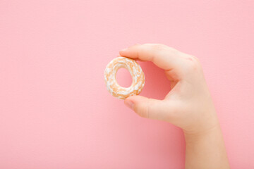 Baby girl fingers holding hard sweet dry bread ring with white sugar glaze on light pink table background. Pastel color. Closeup. Children sweet snacks. Top down view.