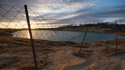 Barbed wire and fence against backdrop of majestic sunset and lake: quarry, sand, picturesque sky.