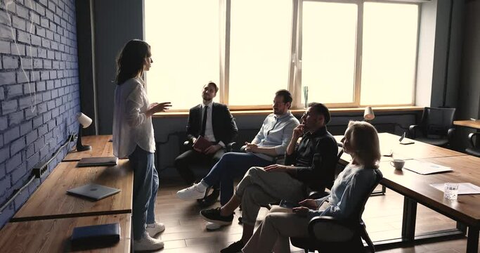 Young business startup leader woman talking to team of colleagues at office meeting, speaking to coworkers sitting in line, asking, answering questions, presenting creative ideas, plan, strategy