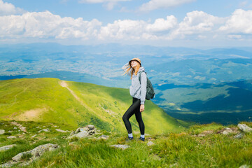 Naklejka na ściany i meble Portrait of a happy woman hiker standing on the slope of mountain ridge against mountains, blue cloudy sky on background. The woman is happy and looking at camera. photo