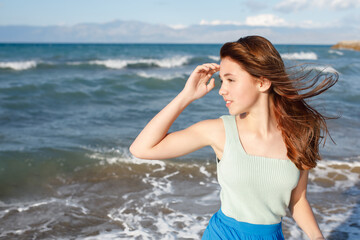 A young girl dressed in bright clothes in the color of the sea looks to the side, her hair flutters in the wind, the background of a beautiful blue sea with waves behind