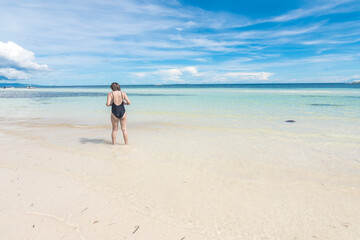 An anonymous female tourist in a black one piece swimsuit takes a casual stroll along a pristine white sand beach.