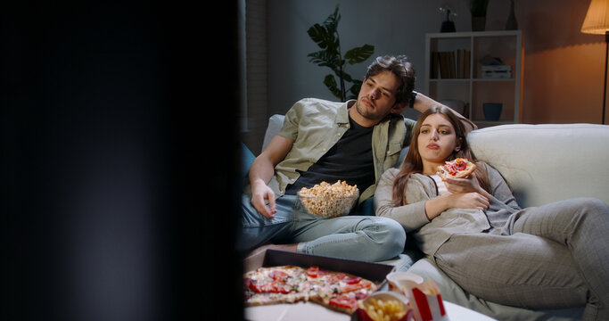 Copy space. Young student caucasian couple spending time together on couch, sitting in front of tv, watching random shows in absolute boredom, tired after a long day eating unhealthy food 