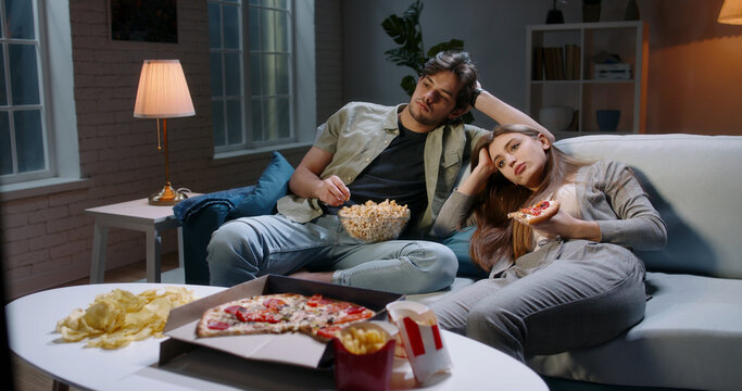 Caucasian Extremely Frustrated Couple Sitting In Front Of Tv Watching Shows And Eating Pizza In Absolute Boredom. Student Couple Tired After A Long Day 