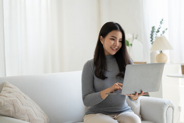 Fototapeta premium Young beautiful Asian woman using laptop computer while sitting on the sofa at her living room.
