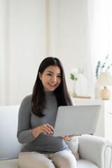 Young beautiful Asian woman using laptop computer while sitting on the sofa at her living room.