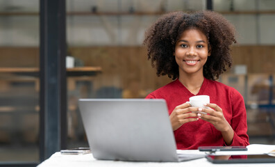 Portrait of smiling black woman sitting at desk, holding coffee cup and using laptop, looking at...