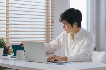 Fototapeta premium Happy and relaxed millennial Asian businessman is working remotely from his home office with his laptop.