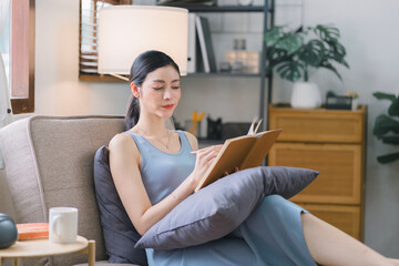 A beautiful, young, smiling and cheerful woman is reading a book while sitting on a couch in the living room.