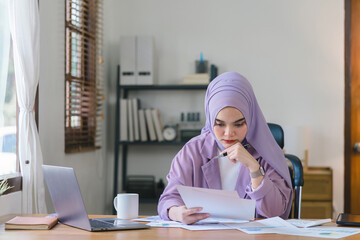 The Muslim millennial woman wearing a purple hijab appears stressed while working on her laptop from home, looking overwhelmed and exhausted.