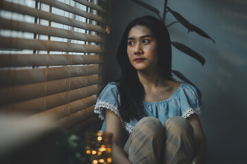 Sad and depressed young woman sitting nere window in the living room, looking outside with a sad...