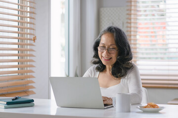 At her home office, the senior entrepreneur wears stylish glasses while working on her computer and taking notes with pen and paper.