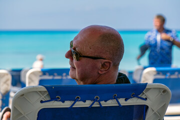A mature gay man sitting on a beach chair relaxing on a tropical beach looks back over his left...