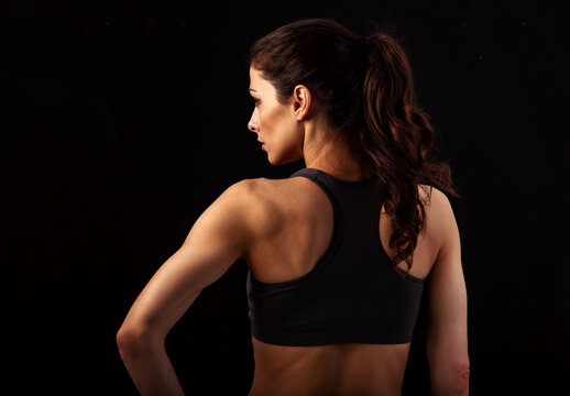 Serious female sporty muscular with ponytail doing stretching workout the shoulders, blades and arms in sport bra, standing on black background with empty space. Back view, profile face