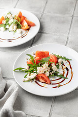 Healthy vegetable salad with fresh arugula, tomato, feta cheese on white plate served for two, gray tile background. Diet menu. Appetizer salad