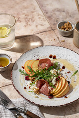 Salad of arugula and prosciutto or ham , sliced apple, cheese, walnuts on rustic plate in composition with cutlery and water glass on pink tile background. Copy space