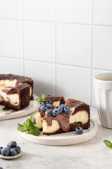 Two pieces of homemade chocolate cheesecake with blueberries, chocolate sauce and mint on a plate with coffee on white kitchen table for morning breakfast. Text space, copy space