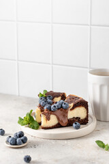 Two pieces of homemade chocolate cheesecake with blueberries, chocolate sauce and mint on a plate with coffee on white kitchen table for morning breakfast. Text space, copy space