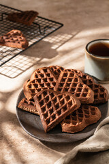 Heart form chocolate waffle cookies in brown ceramic plate with tea cup on beige background. Sunny morning breakfast with hursh shadows