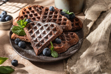 Heart form chocolate waffle cookies in ceramic plate decorated with berries, powdered sugar and mint with tea on beige background. Sunny morning breakfast with hursh shadows