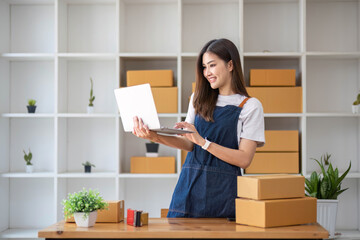 Fototapeta na wymiar A portrait of a young Asian woman, e-commerce employee sitting in the office full of packages in the background write note of orders and a calculator, for SME business ecommerce and delivery business.