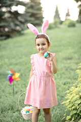 Little cute girl wear bunny ears holding basket with colorful painted eggs on Easter egg hunt in park