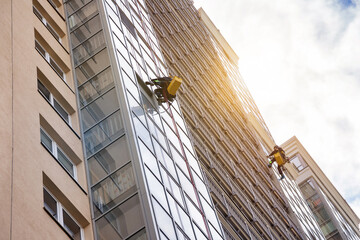 Professional industrial mountaineer workers washing exterior facade glazing, hangs over building,...