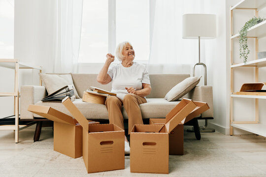 Elderly Woman Sits On A Sofa At Home With Boxes. Collecting Things With Memories And Moving And Happiness Smile.