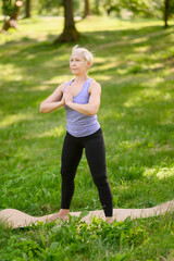 Fototapeta premium Middle-aged woman doing yoga in sportswear outdoors in a city park in Namaskarasana - Seated Prayer Pose. Concept of stretching, pilates, healthy lifestyle.