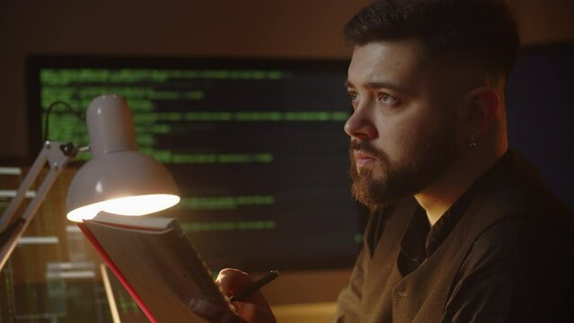 The software developer writes notes in his notebook. Close-up of a male developer sitting in front of a monitor with running lines of program code.