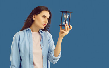 Beautiful, young brunette woman, dressed in light T-shirt and light blue oversized shirt, stares intently at the hourglass in her hand. Portrait on isolated blue background in the studio. - obrazy, fototapety, plakaty
