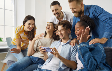 Cheerful friends watching something very funny on mobile phone. Group of happy young diverse people hanging out, having fun, sitting on sofa and laughing at hilarious meme or joke on social media