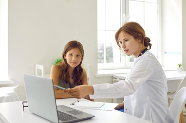 Obraz na płótnie Canvas Young professional woman doctor with serious consulting a teen girl on checkup appointment showing something in laptop screen. Pediatrician consultation visit in clinic. Healthcare, medicine concept.