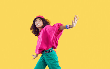 Happy child dancing. Cheerful little African American girl wearing loose oversized fuschia top and green jeans with trendy eyelet belt dancing isolated on solid yellow background. Kids fashion concept - obrazy, fototapety, plakaty