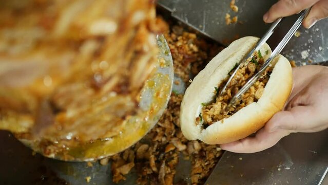 Top view chef's hand holding a cut bun puts juicy pieces of fried meat inside using serving tongs on a rotating vertical grill in a fast food restaurant. Cooking doner kebab in bread. Fast food