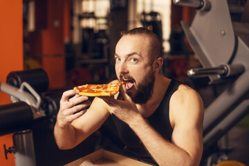 A young man with a beard is happy to eat pizza in the gym.