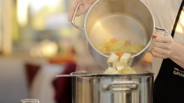 The cook strains the broth with potatoes, celery root and carrots through a sieve. a cook in a restaurant kitchen pours liquid from a broth of potatoes, celery and carrots from one pan to another