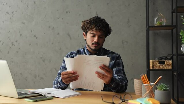 Young satisfied confident successful employee IT Indian business man wear casual clothes shirt sit work at office desk with laptop pc computer read check paper documents Professional staff job concept