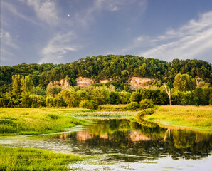 Beautiful view of Midwestern river bluffs with ponds in front of them and rising moon on summer evening; bluffs, trees and  flowers reflect in  calm water