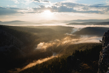 Fog creeps into the river valley early summer morning, sunrise in the mountains, rock cliff, sun glare, golden hour in photography, nature morning