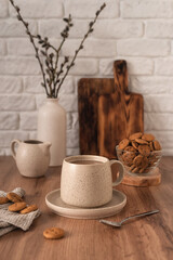 A ceramic cup with coffee, a milk jug, a glass bowl with cookies and a vase with branches on a wooden kitchen table. Still life in the style of Scandi. Home comfort.